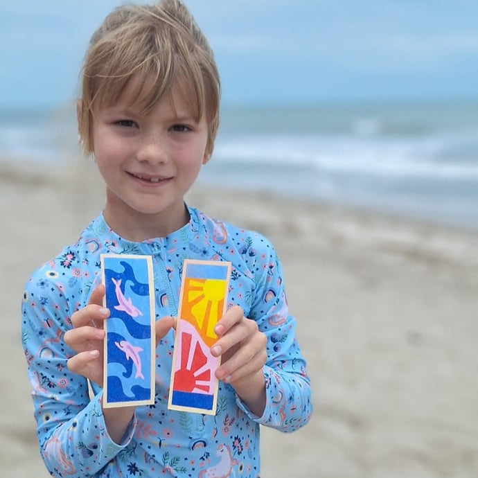 Niña sonriente mostrando marcadores de libros creados con arena de colores SandArt en la playa, actividad creativa y portátil para vacaciones que fomenta la expresión artística